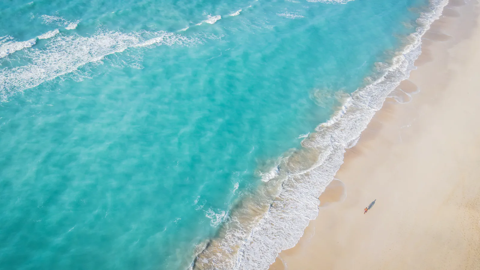 Pogled iz zraka na turkizne valove ob peščeni obali otoka Saadiyat; na plaži je majhna oseba.