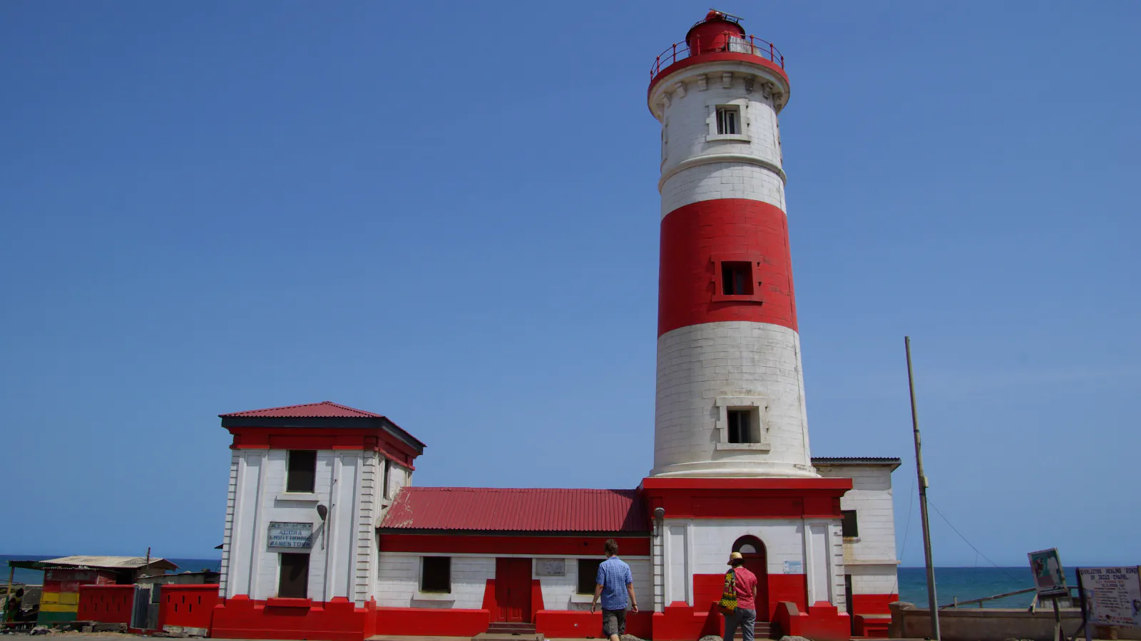 A white lighthouse tower with red bands and a red lantern rising behind a white and red building, with blue sky and the sea beyond.
