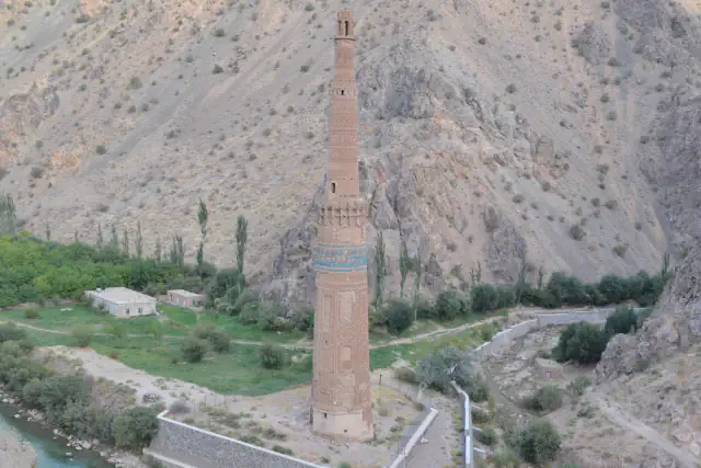 Visok opečni minaret z modrim ploščičnim pasom stoji ob turkizni reki v ozki gorski dolini; za njim se dvigajo gola skalnata pobočja, okoli njegovega podnožja pa je kamnit podporni zid