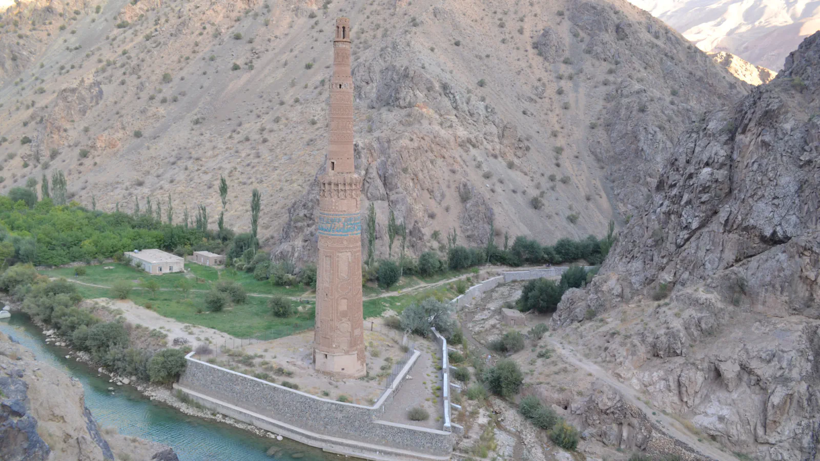 A tall brick minaret with a blue tiled band stands beside a turquoise river in a narrow mountain valley, with bare rocky slopes behind it and a stone retaining wall around its base