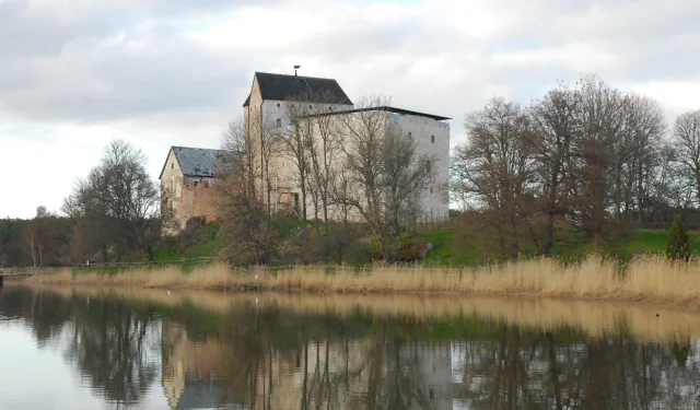 Kastelholm Castle seen across a reed-edged bay: pale stone buildings with dark roofs on a low green rise among bare trees, mirrored in calm water under a cloudy sky