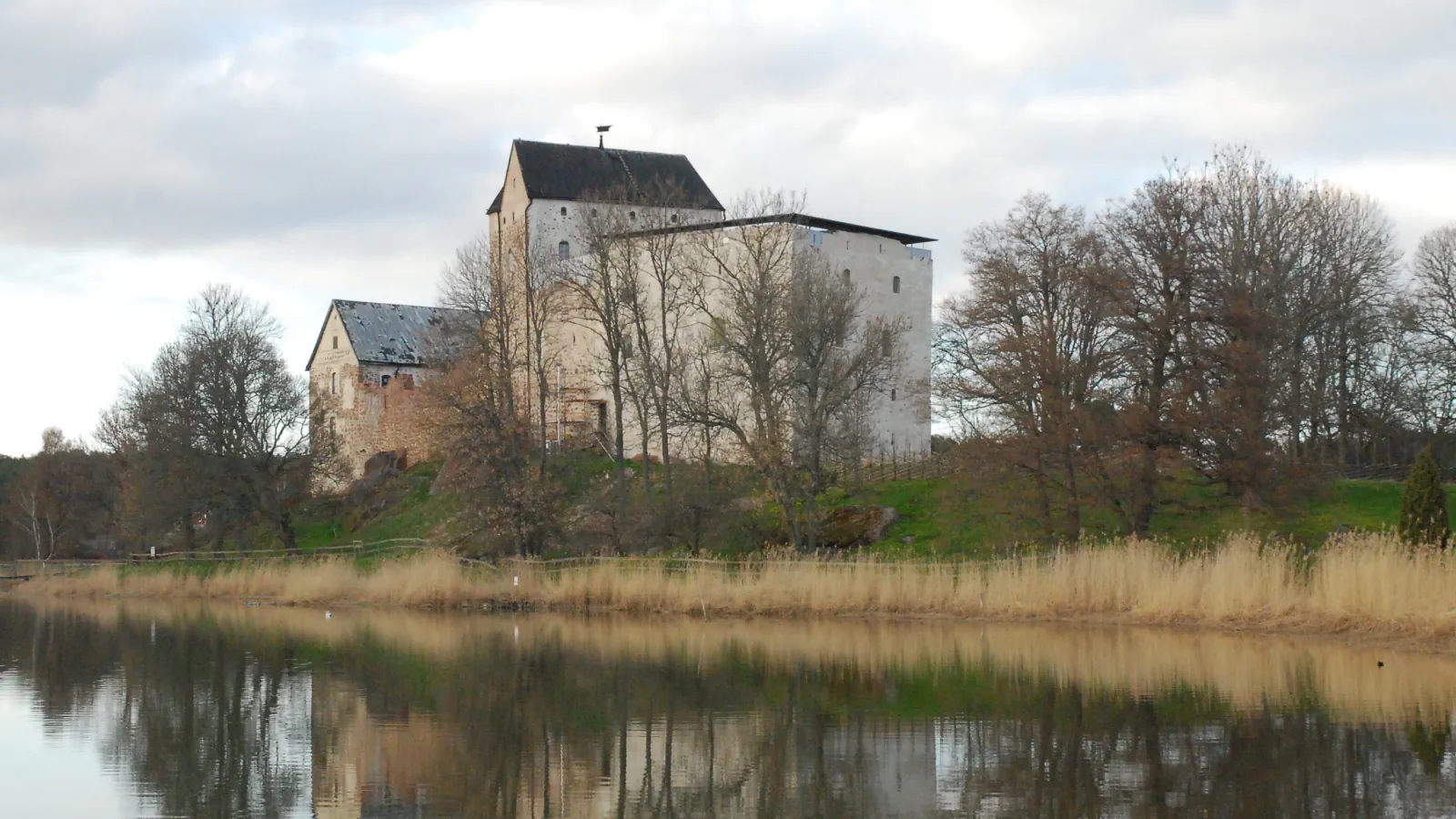 Kastelholm Castle seen across a reed-edged bay: pale stone buildings with dark roofs on a low green rise among bare trees, mirrored in calm water under a cloudy sky