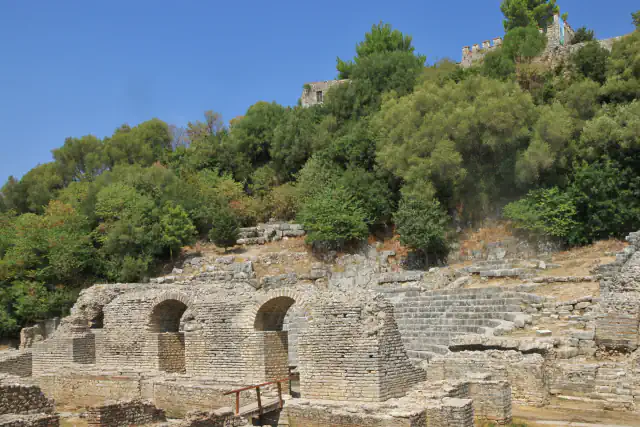 Stone ruins of the theatre at Butrint among trees, with the walls of a fortress on the hill above