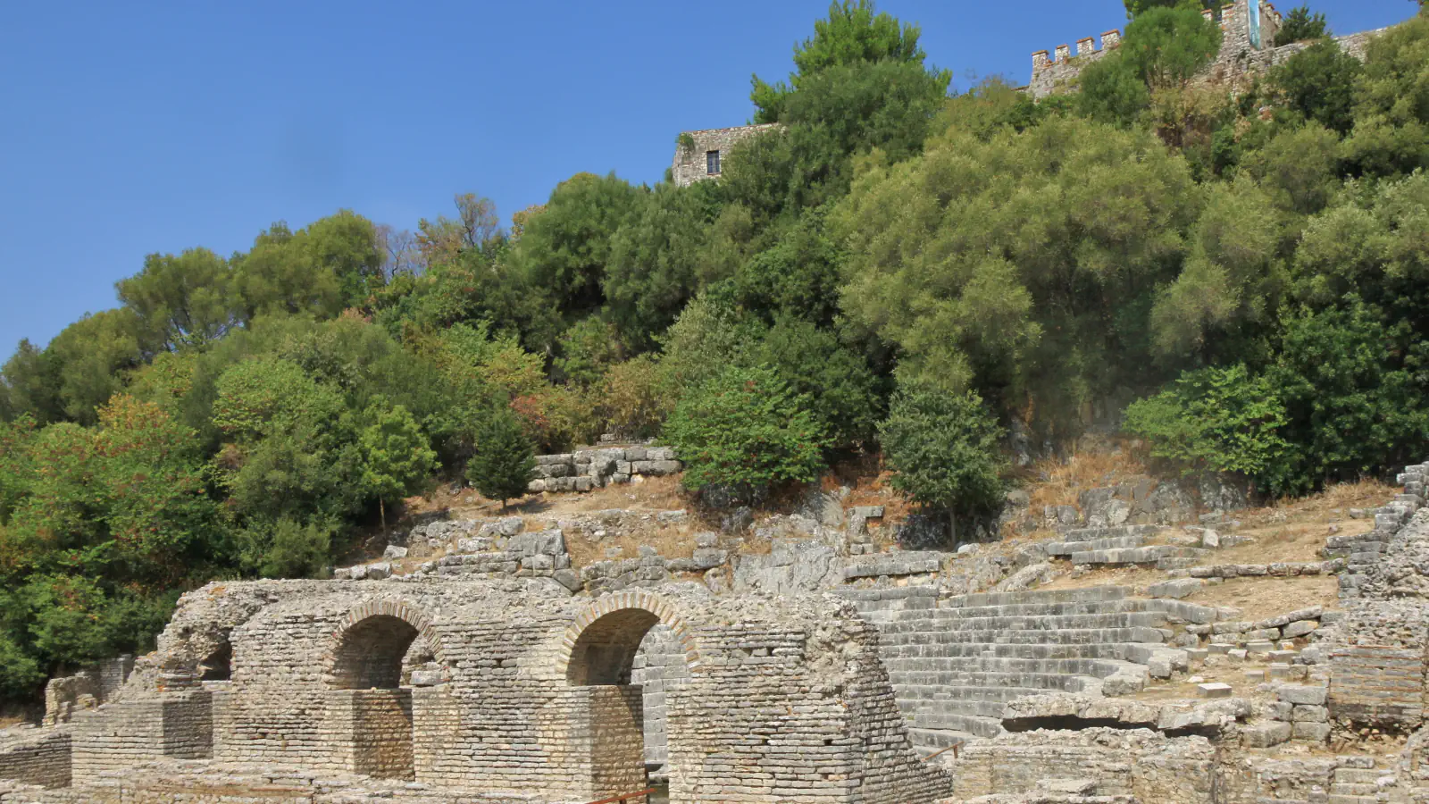 Stone ruins of the theatre at Butrint among trees, with the walls of a fortress on the hill above