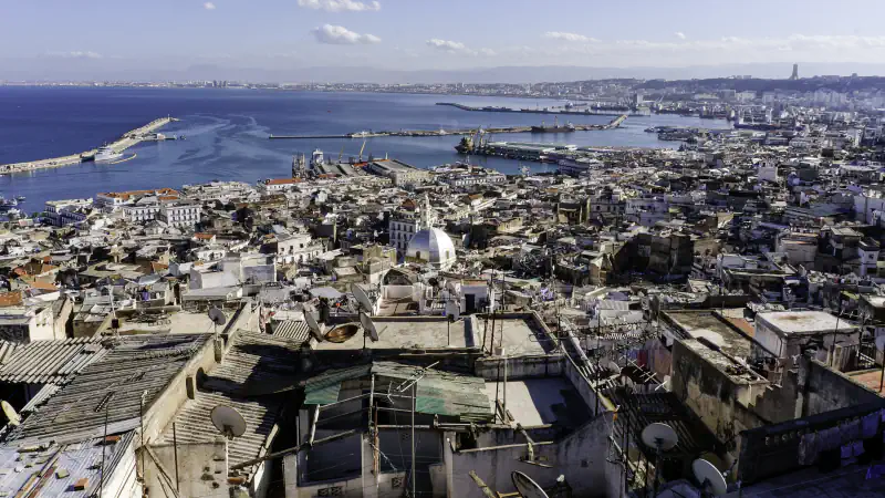 Flat roof terraces of the Casbah in the foreground, a white dome among the dense houses of the lower town, and the harbour moles and the Bay of Algiers beyond