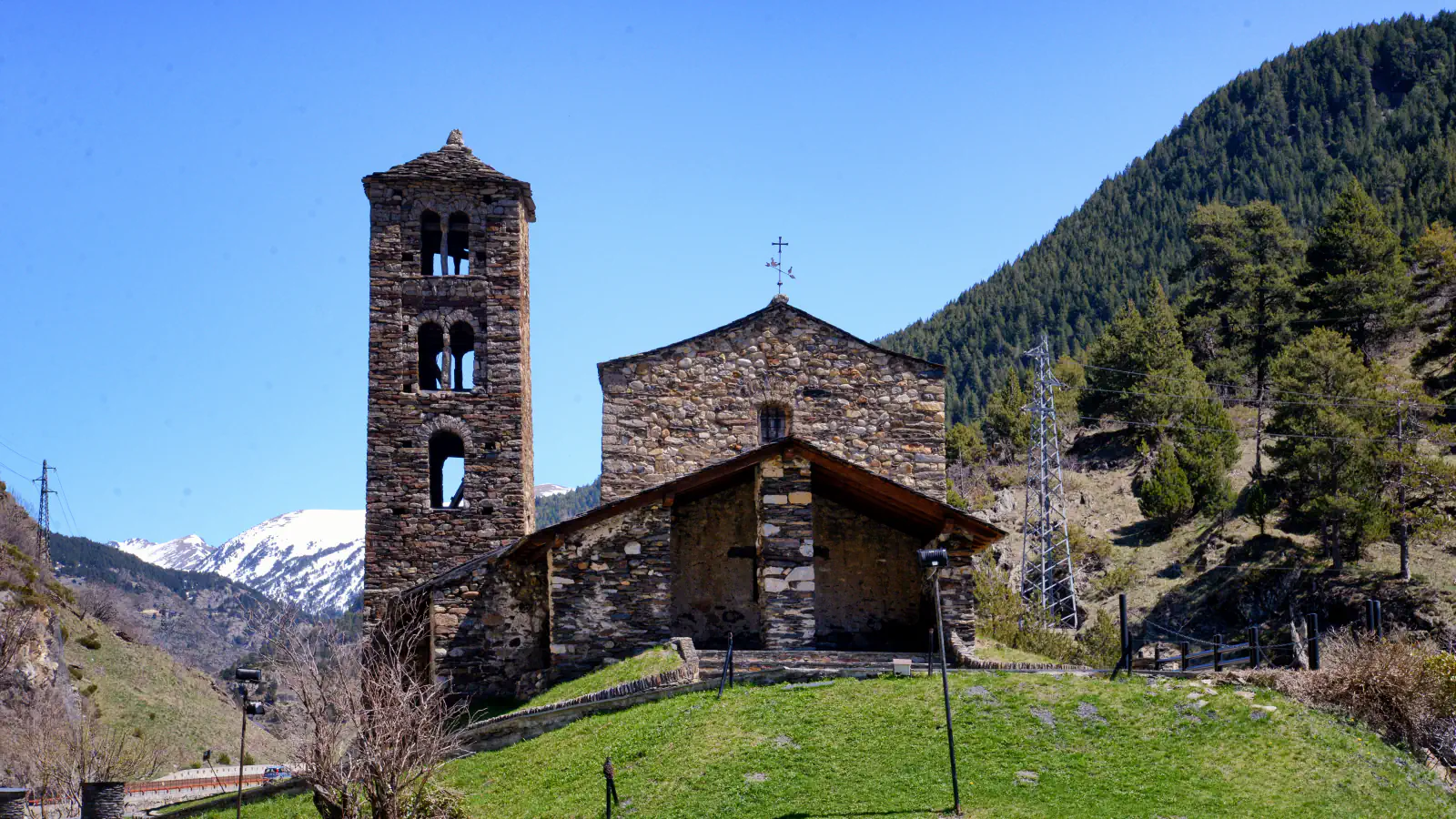 A grey-stone Romanesque church with a tall square bell tower with arched openings at left and a porch at the front stands on a grassy mound, with a forested mountainside to the right, a snow-capped peak behind and a clear blue sky