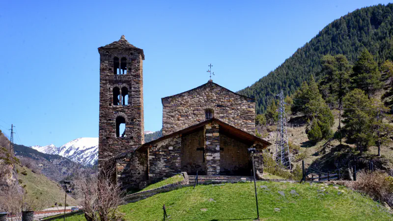 A grey-stone Romanesque church with a tall square bell tower with arched openings at left and a porch at the front stands on a grassy mound, with a forested mountainside to the right, a snow-capped peak behind and a clear blue sky