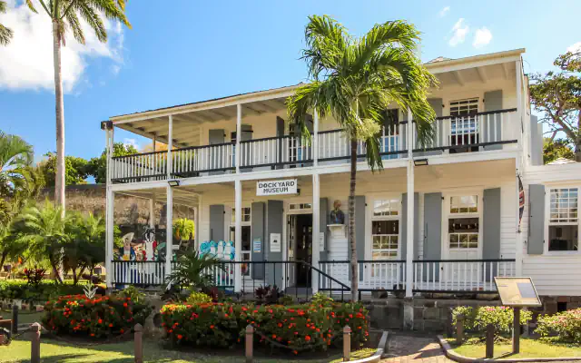 A white two-storey wooden building with covered verandas, grey shutters and a sign reading Dockyard Museum, with a palm tree in front, red flowering shrubs below and a bright blue sky
