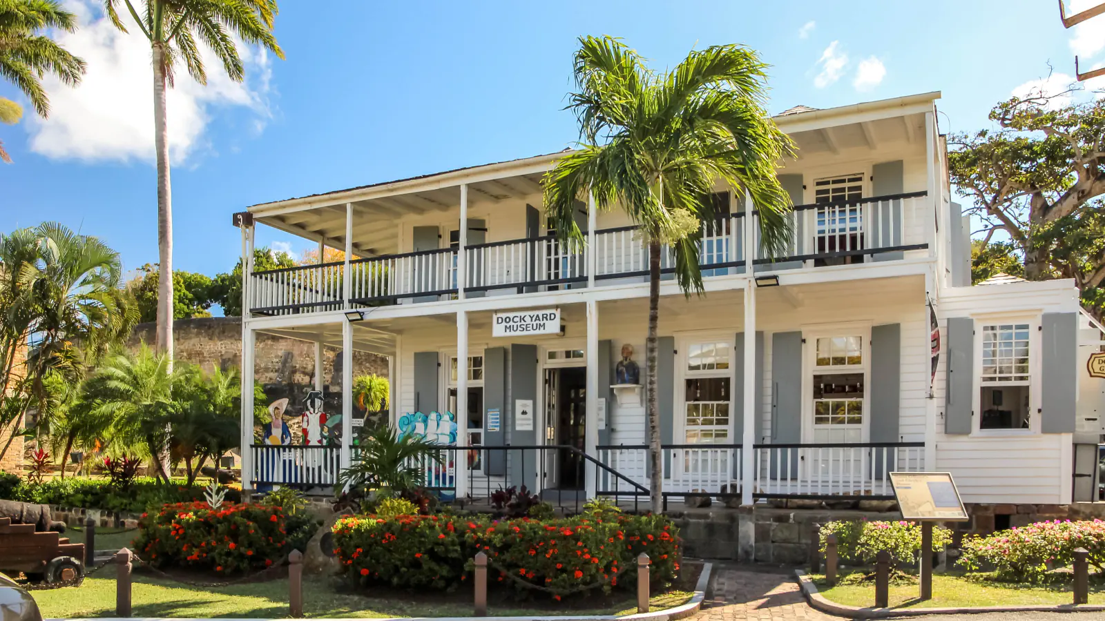 A white two-storey wooden building with covered verandas, grey shutters and a sign reading Dockyard Museum, with a palm tree in front, red flowering shrubs below and a bright blue sky