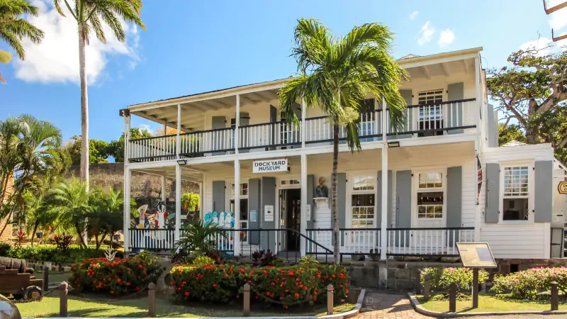 A white two-storey wooden building with covered verandas, grey shutters and a sign reading Dockyard Museum, with a palm tree in front, red flowering shrubs below and a bright blue sky