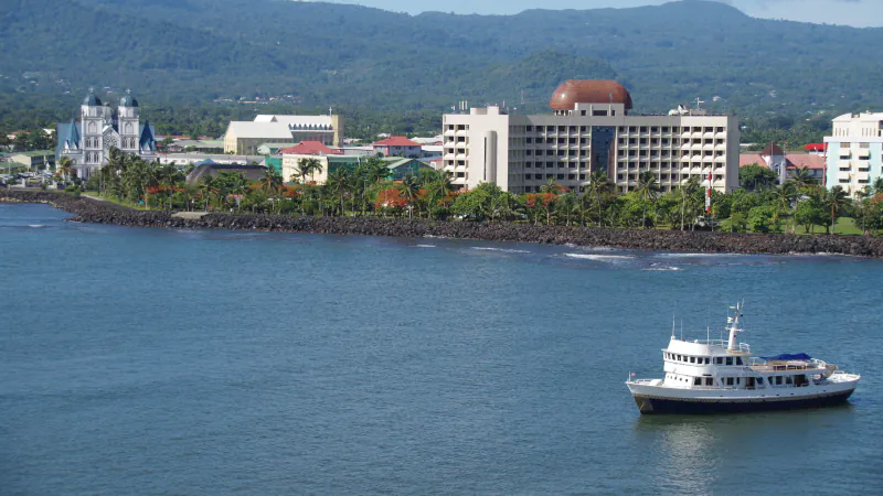Apia’s waterfront with a cathedral and a large multi-storey building, seen from the harbour with a boat in the foreground.