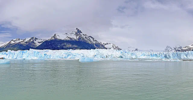 A wall of blue-white ice at the front of a glacier rising from grey-green lake water, with snow-capped mountains and low cloud behind