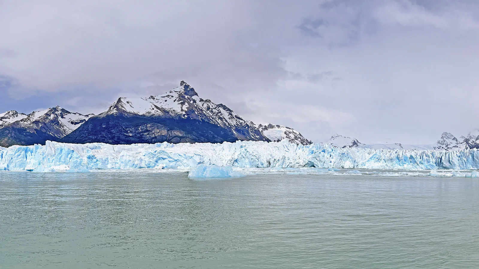 A wall of blue-white ice at the front of a glacier rising from grey-green lake water, with snow-capped mountains and low cloud behind