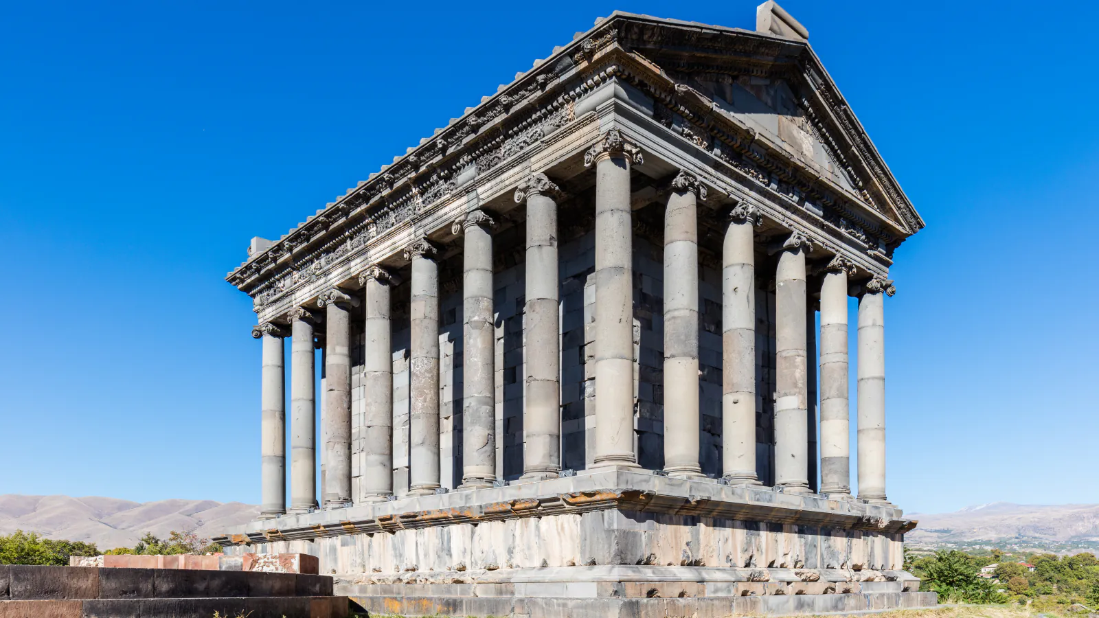 Ionic columns and pediment of the Garni temple under a clear blue sky, with hills behind
