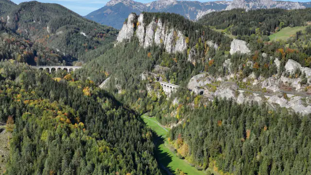 Aerial view of the Semmering Railway’s stone viaducts and tunnel portals set into forested slopes, with the Rax massif on the horizon