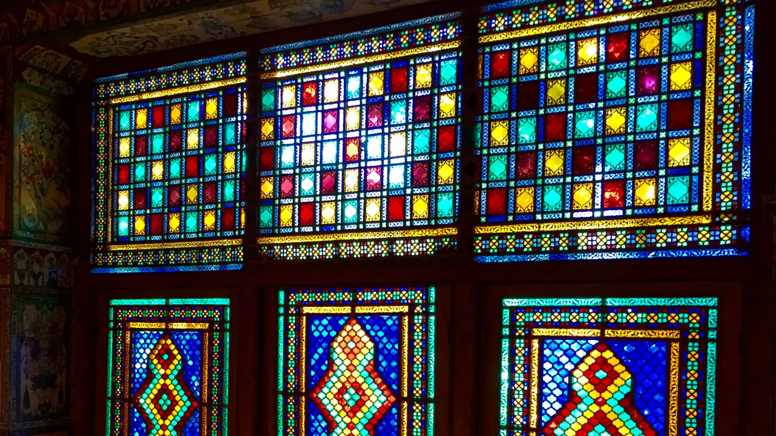 Six wooden-framed windows glow with small panes of red, yellow, turquoise and deep blue glass, the upper three in square grids and the lower three with diamond-shaped medallions at their centre