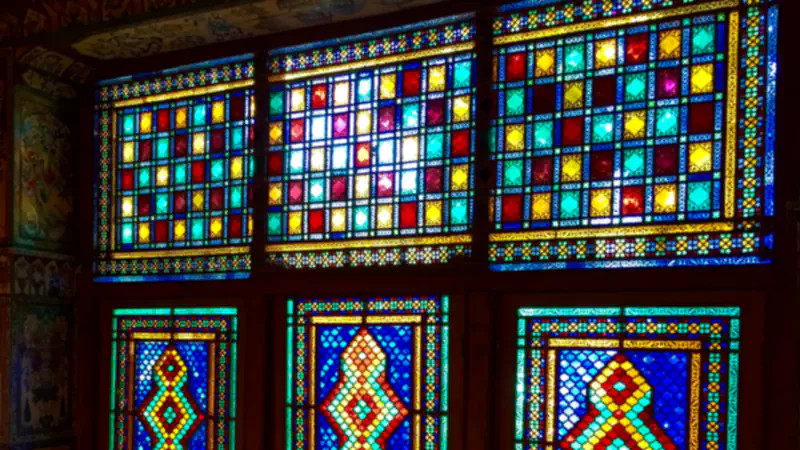 Six wooden-framed windows glow with small panes of red, yellow, turquoise and deep blue glass, the upper three in square grids and the lower three with diamond-shaped medallions at their centre