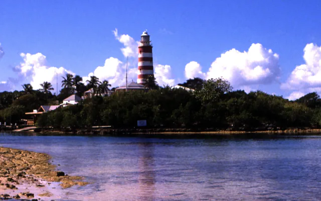 Red-and-white striped lighthouse above a tree-lined Hope Town harbour, with low buildings along the shore and clouds in a blue sky