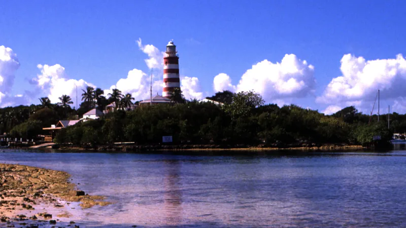 Red-and-white striped lighthouse above a tree-lined Hope Town harbour, with low buildings along the shore and clouds in a blue sky