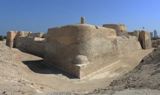 A sand-coloured stone fort with rounded corner bastions and a small domed turret rises behind a dry ditch under a clear blue sky, with a few distant tall buildings on the right