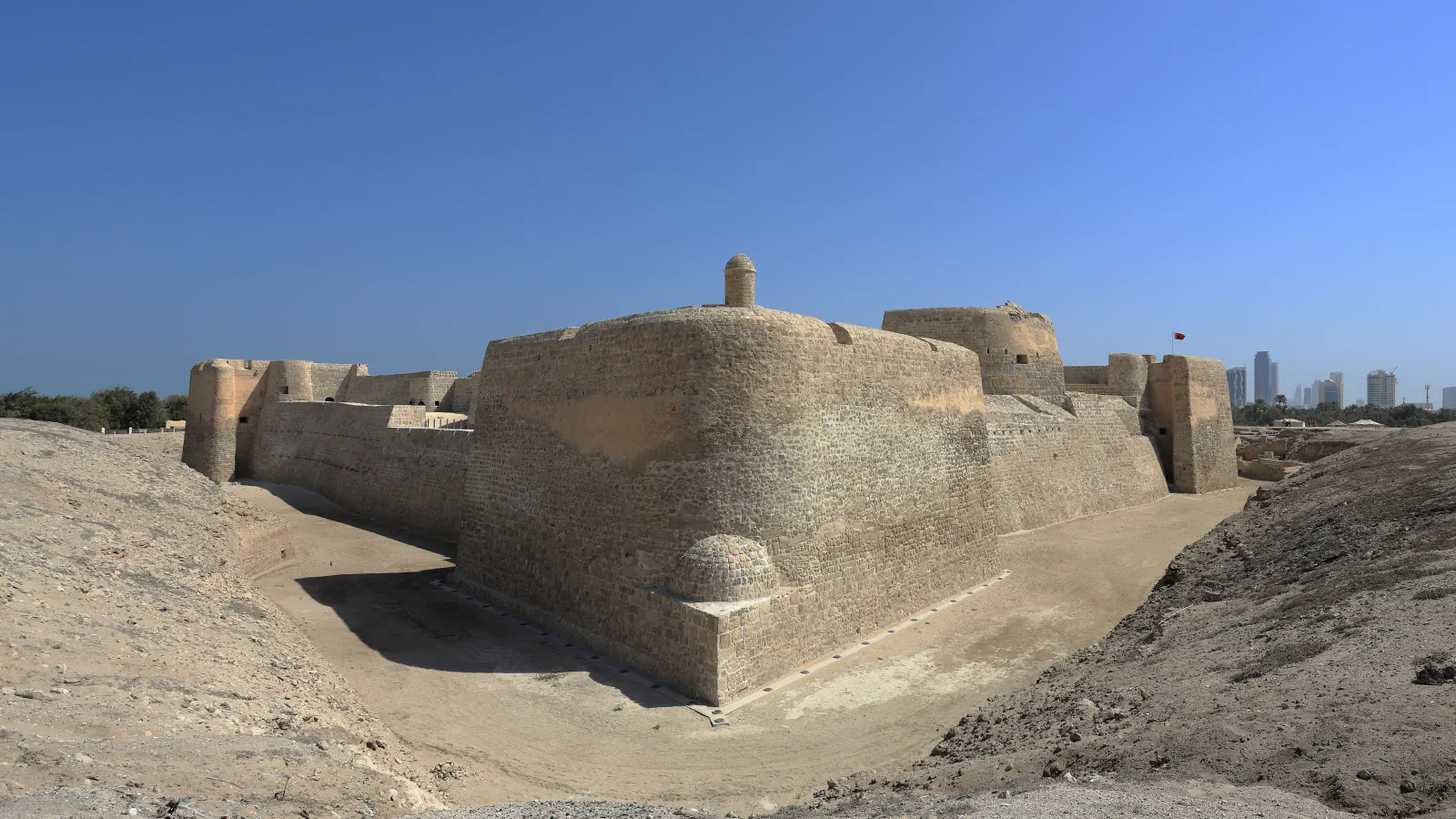 A sand-coloured stone fort with rounded corner bastions and a small domed turret rises behind a dry ditch under a clear blue sky, with a few distant tall buildings on the right