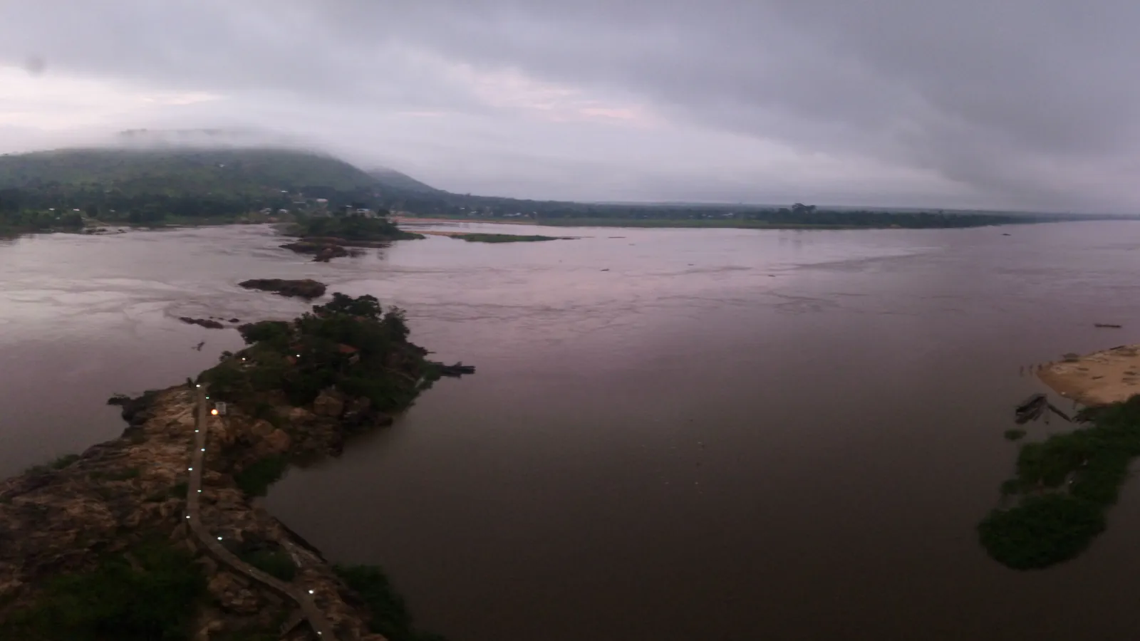 The broad brown Oubangui River under heavy grey cloud, with a rocky, tree-covered island and a footpath in the foreground and a low green hill on the far bank