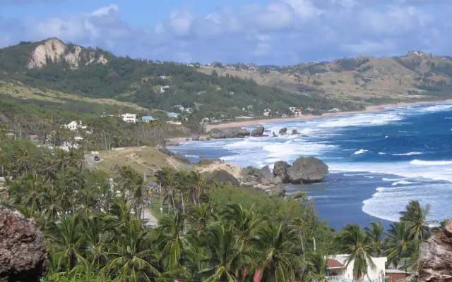 Waves break around large coastal rocks at Bathsheba, with palm trees and green hills along the east coast of Barbados.