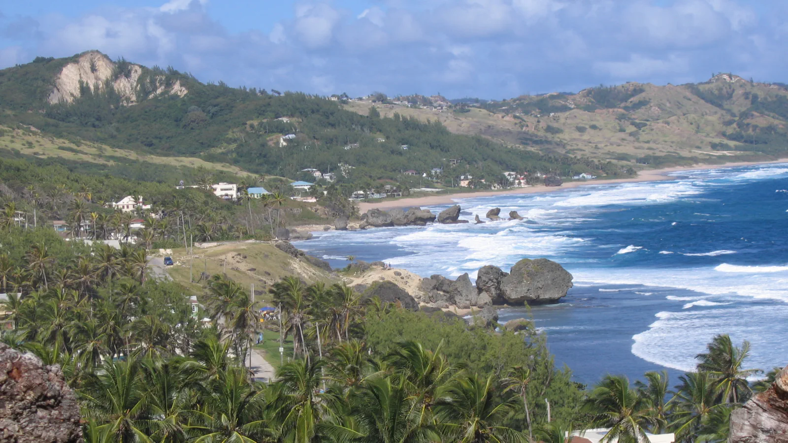 Waves break around large coastal rocks at Bathsheba, with palm trees and green hills along the east coast of Barbados.
