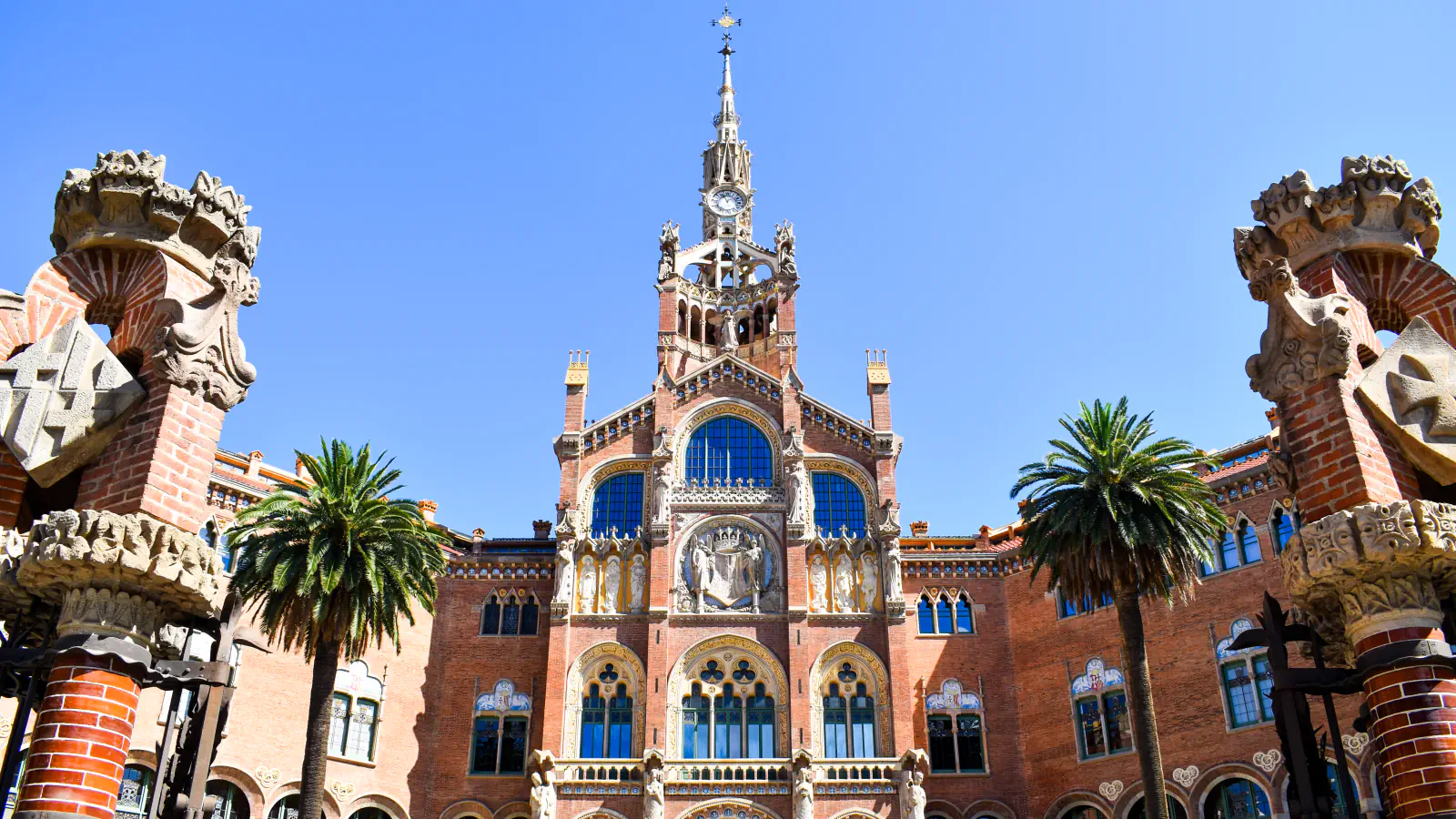Brick façade of the Hospital de Sant Pau with its clock tower and spire, framed by two ornate brick pillars and palm trees