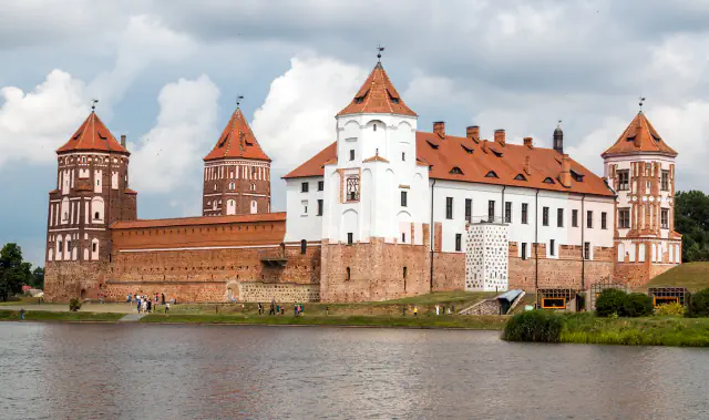 A red-brick castle with whitewashed upper floors and several towers under pointed tile roofs stands behind a stretch of water under a cloudy sky