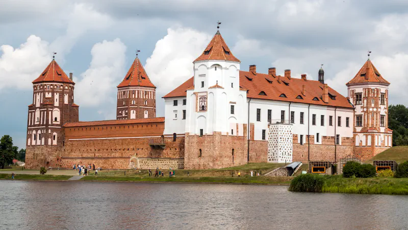 A red-brick castle with whitewashed upper floors and several towers under pointed tile roofs stands behind a stretch of water under a cloudy sky