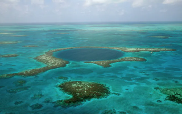 The Great Blue Hole’s dark circular sinkhole, encircled by pale reef and turquoise shallows, with a small sailboat near the outer rim