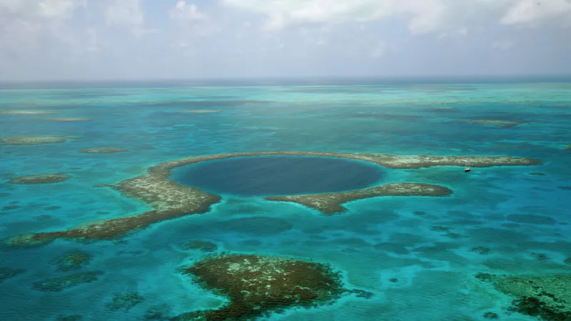 The Great Blue Hole’s dark circular sinkhole, encircled by pale reef and turquoise shallows, with a small sailboat near the outer rim