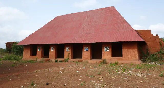 A low earthen palace with a steep red corrugated-metal roof, open bays and small coloured relief panels in its ochre walls, standing on bare red ground beneath a pale sky