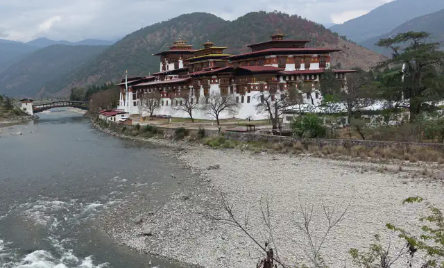 A river runs beside a white-walled dzong with red-roofed, timber-framed upper storeys on the right, a wooden bridge upstream and forested hills behind under a cloudy sky