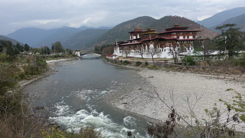 A river runs beside a white-walled dzong with red-roofed, timber-framed upper storeys on the right, a wooden bridge upstream and forested hills behind under a cloudy sky