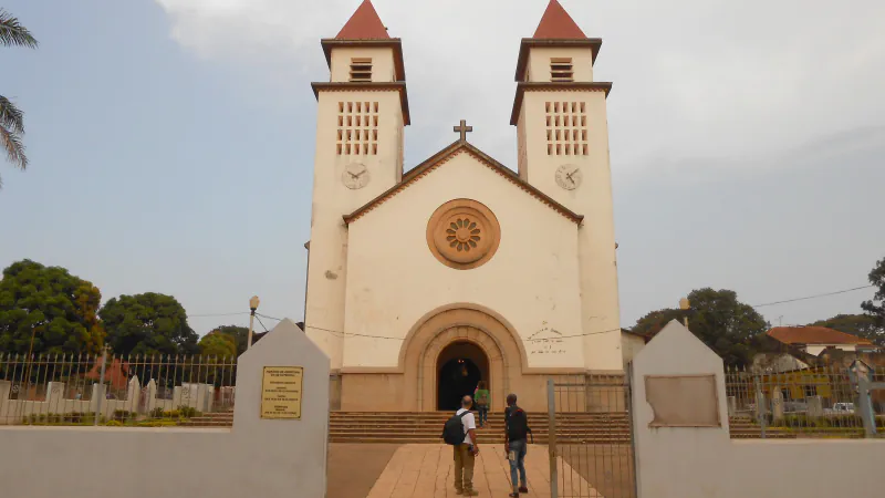 Front of Bissau Cathedral: a cream-coloured façade with a round window and an arched doorway between two square towers with red pointed roofs and clock faces, with two people at the foot of the steps