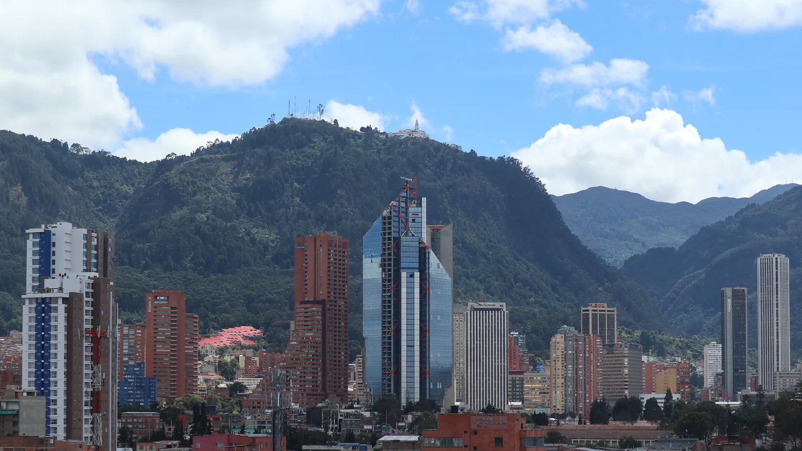 Tall glass and brick towers of central Bogotá in front of the forested Monserrate ridge, under a blue sky with large white clouds