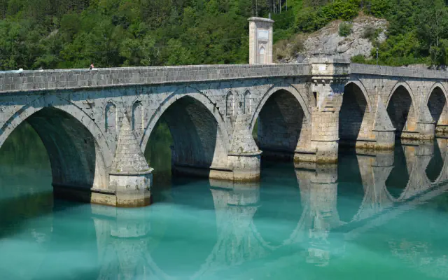 Kamniti oboki mostu Mehmed Paše Sokolovića nad bledo zeleno Drino, s koničastimi glavami stebrov; oboki se zrcalijo v vodi