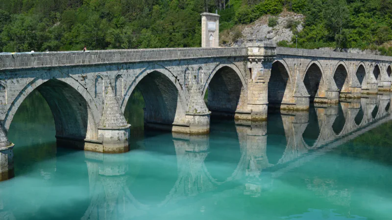 Stone arches of the Mehmed Paša Sokolović Bridge over the pale green Drina, with pointed pier heads and the arches reflected in the water