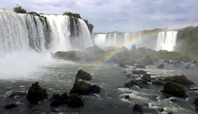 Iguaçu Falls with a rainbow arching over the river, wide curtains of white water at left and dark mossy rocks in the foreground