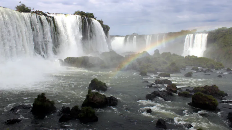 Iguaçu Falls with a rainbow arching over the river, wide curtains of white water at left and dark mossy rocks in the foreground