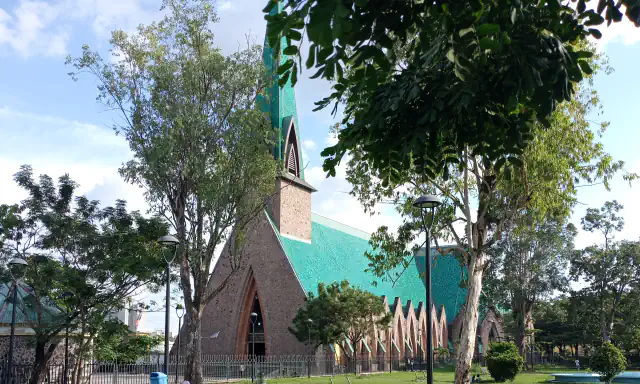 The Sainte-Anne-du-Congo basilica in Brazzaville: a pointed brick gable, a tall green-tiled spire and a long green-tiled roof behind a railing, seen through trees from a lawn with lamp posts
