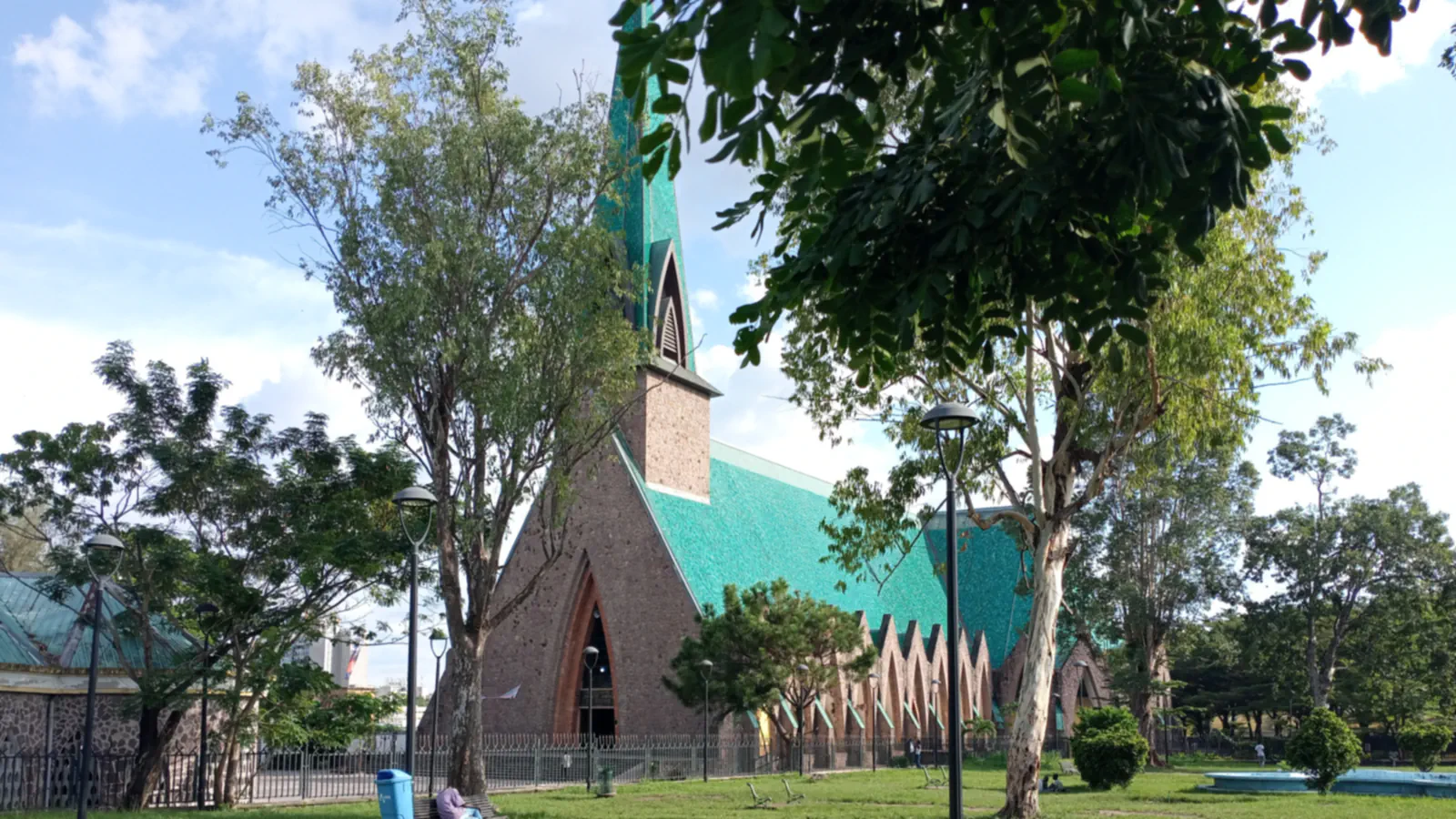 The Sainte-Anne-du-Congo basilica in Brazzaville: a pointed brick gable, a tall green-tiled spire and a long green-tiled roof behind a railing, seen through trees from a lawn with lamp posts