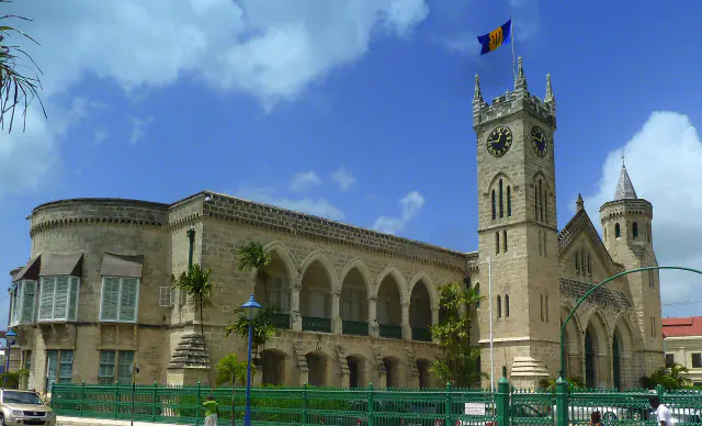 A pale stone Parliament building with a long arcade of pointed arches and a square clock tower flying Barbados’s flag, behind a green iron fence under a blue sky.