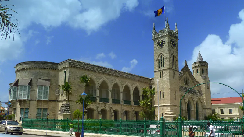 A pale stone Parliament building with a long arcade of pointed arches and a square clock tower flying Barbados’s flag, behind a green iron fence under a blue sky.