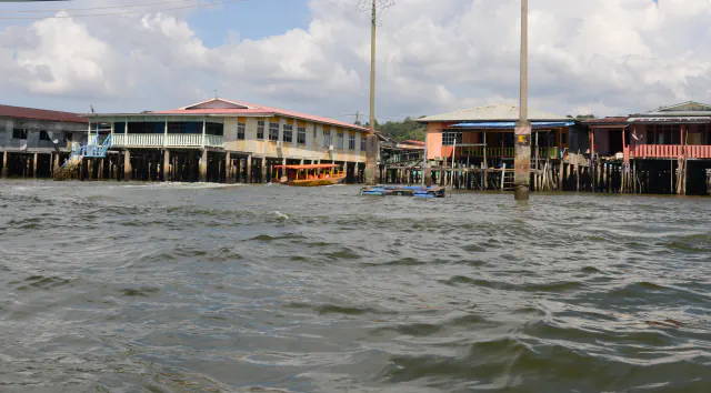 Stilt houses with pastel walls and corrugated roofs stand on dark timber piles beside the Brunei River; a yellow water taxi crosses rippling water.