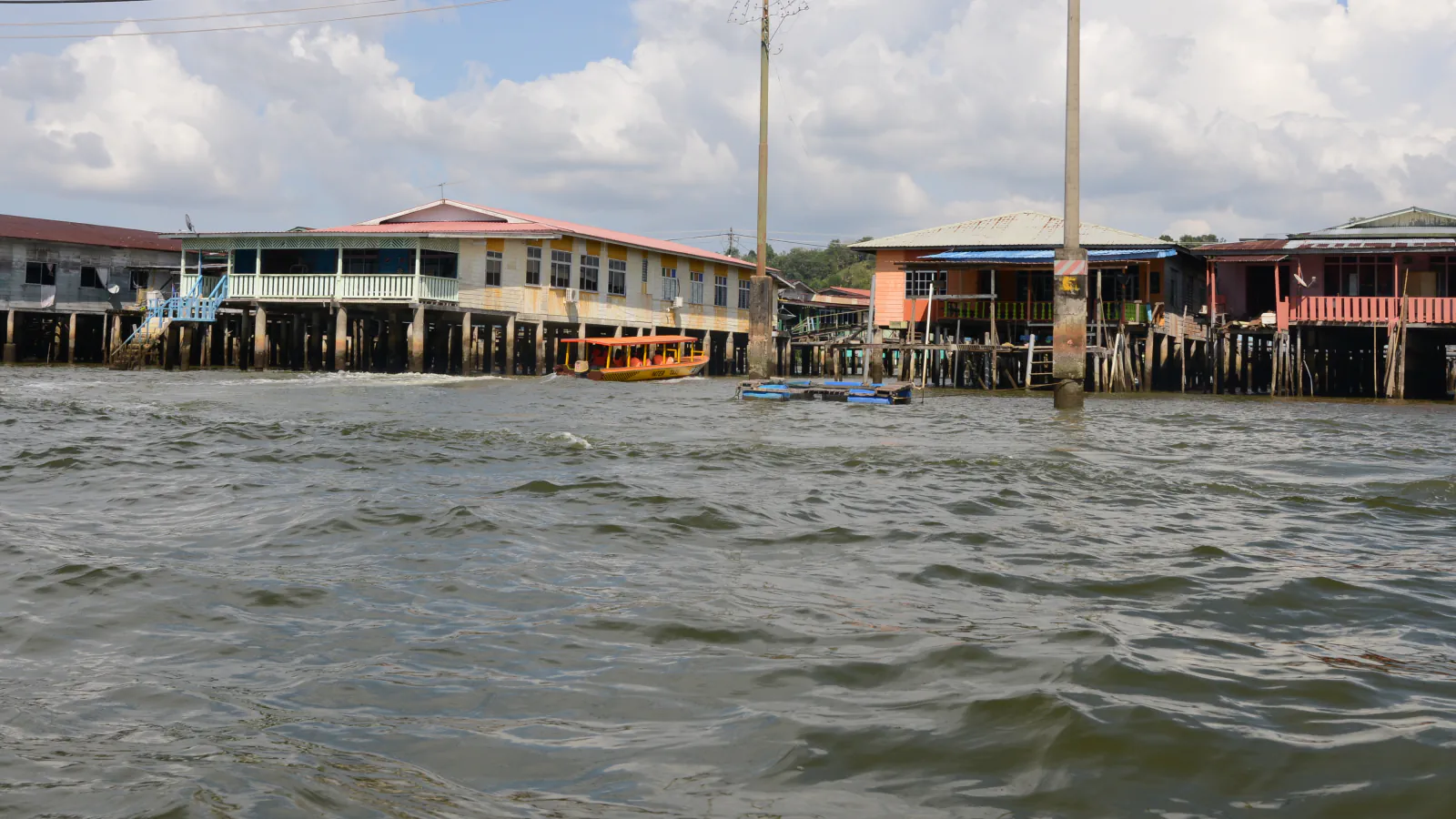 Stilt houses with pastel walls and corrugated roofs stand on dark timber piles beside the Brunei River; a yellow water taxi crosses rippling water.