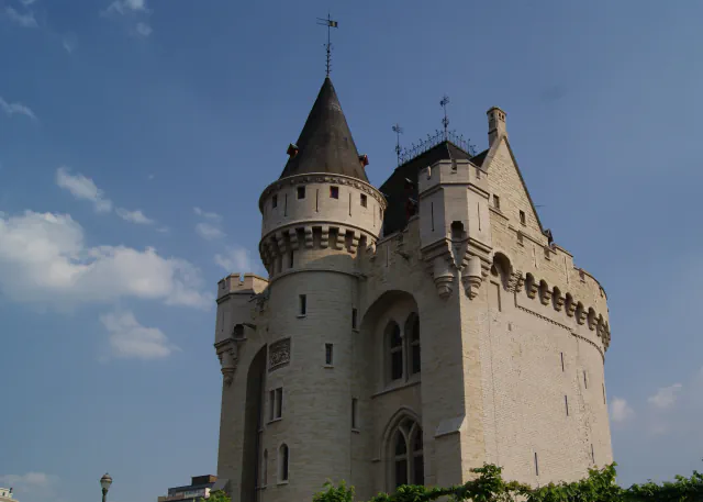 The Halle Gate in Brussels seen from the city side: a pale stone medieval gate with a conical-roofed round tower, battlements and a dark roof, rising above trees against a blue sky