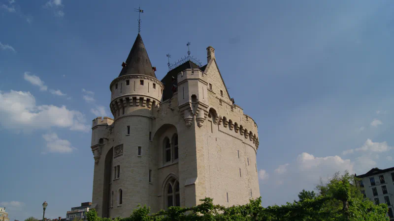 The Halle Gate in Brussels seen from the city side: a pale stone medieval gate with a conical-roofed round tower, battlements and a dark roof, rising above trees against a blue sky